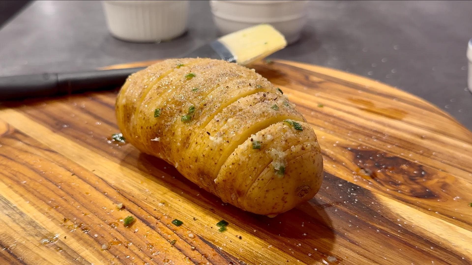 Cooked hasselback potato on wooden cutting board, sliced and seasoned with herbs and butter.
