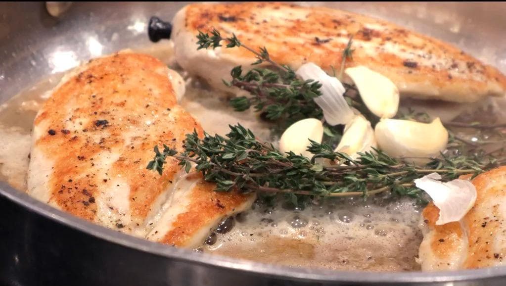 Pan-seared chicken breasts in butter with fresh thyme sprigs and garlic cloves in a stainless steel pan.