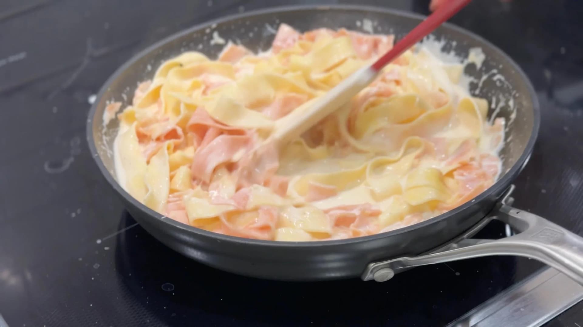 Creamy Alfredo sauce with cooked pasta and diced ham being tossed together in a skillet with a wooden spoon.