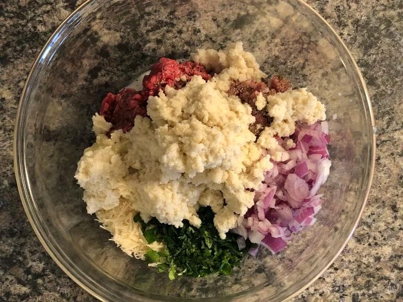 Raw meatball ingredients mixed in a glass bowl: ground beef and pork, breadcrumbs, diced onion, garlic, cheese, and fresh parsley.
