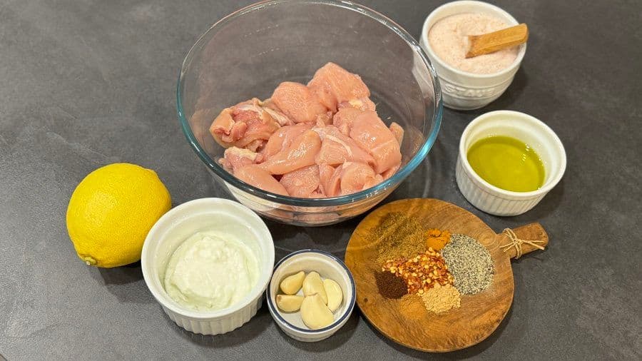 Chicken shawarma ingredients arranged on gray surface: raw chicken pieces, lemon, mayo, olive oil, garlic, and spices on wooden board.