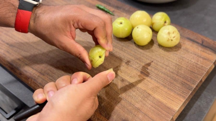 Hands separating amla seeds from flesh on a wooden cutting board with whole green gooseberries nearby.