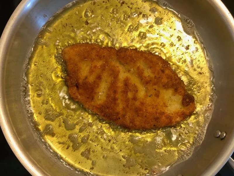 Golden-brown breaded chicken fillet frying in hot oil in a stainless steel pan.