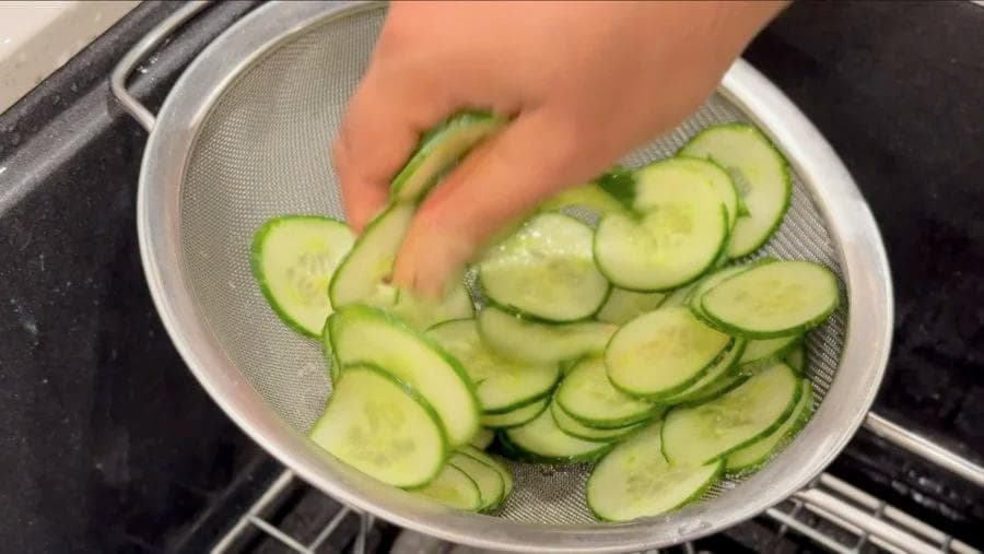 Sliced cucumbers in a sieve being rinsed under water during preparation of German cucumber salad.