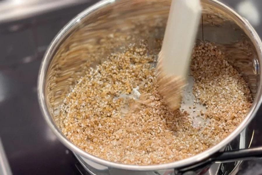 Cracked wheat being roasted in ghee in a saucepan until golden brown, stirred with a wooden spoon.