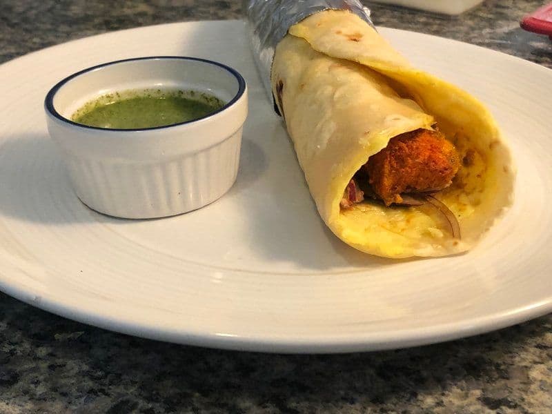 Chicken kathi roll wrapped in parchment paper served with green cilantro chutney on a white plate.