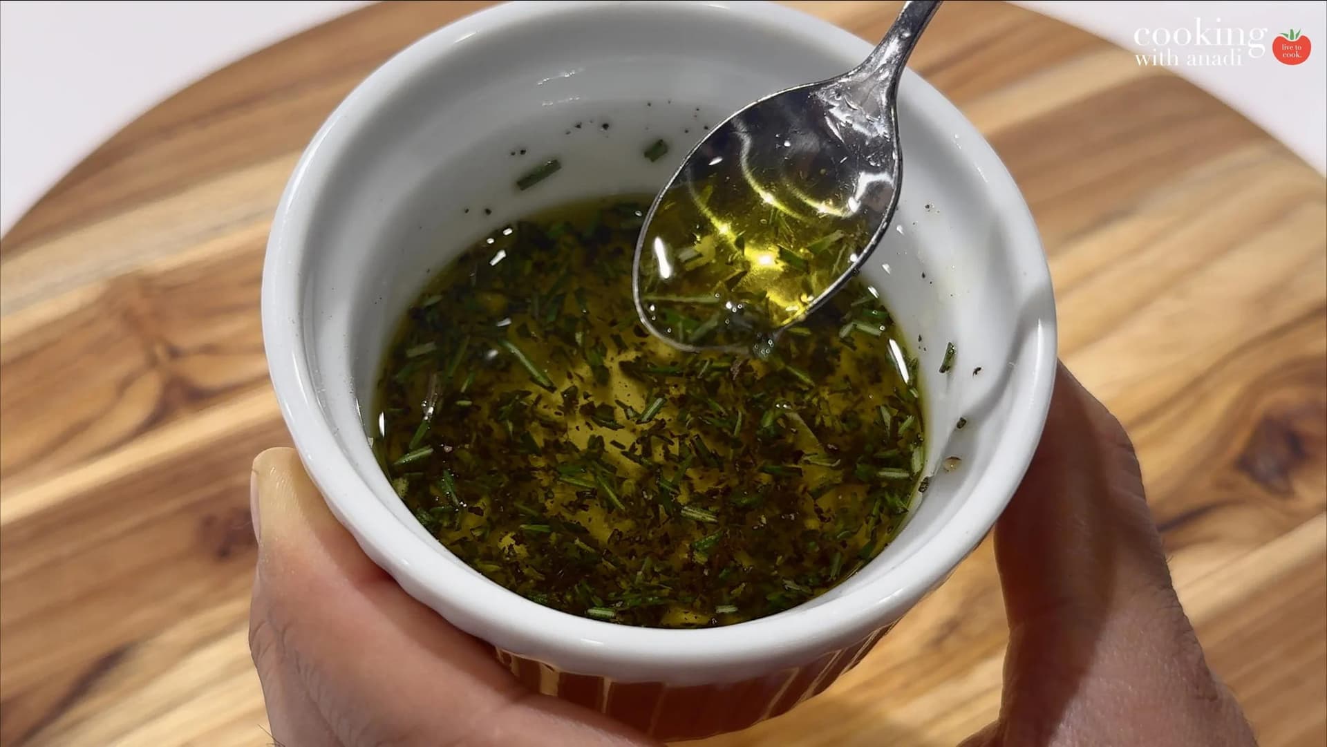 Herb oil with chopped rosemary and thyme in a white ramekin, being stirred with a spoon.