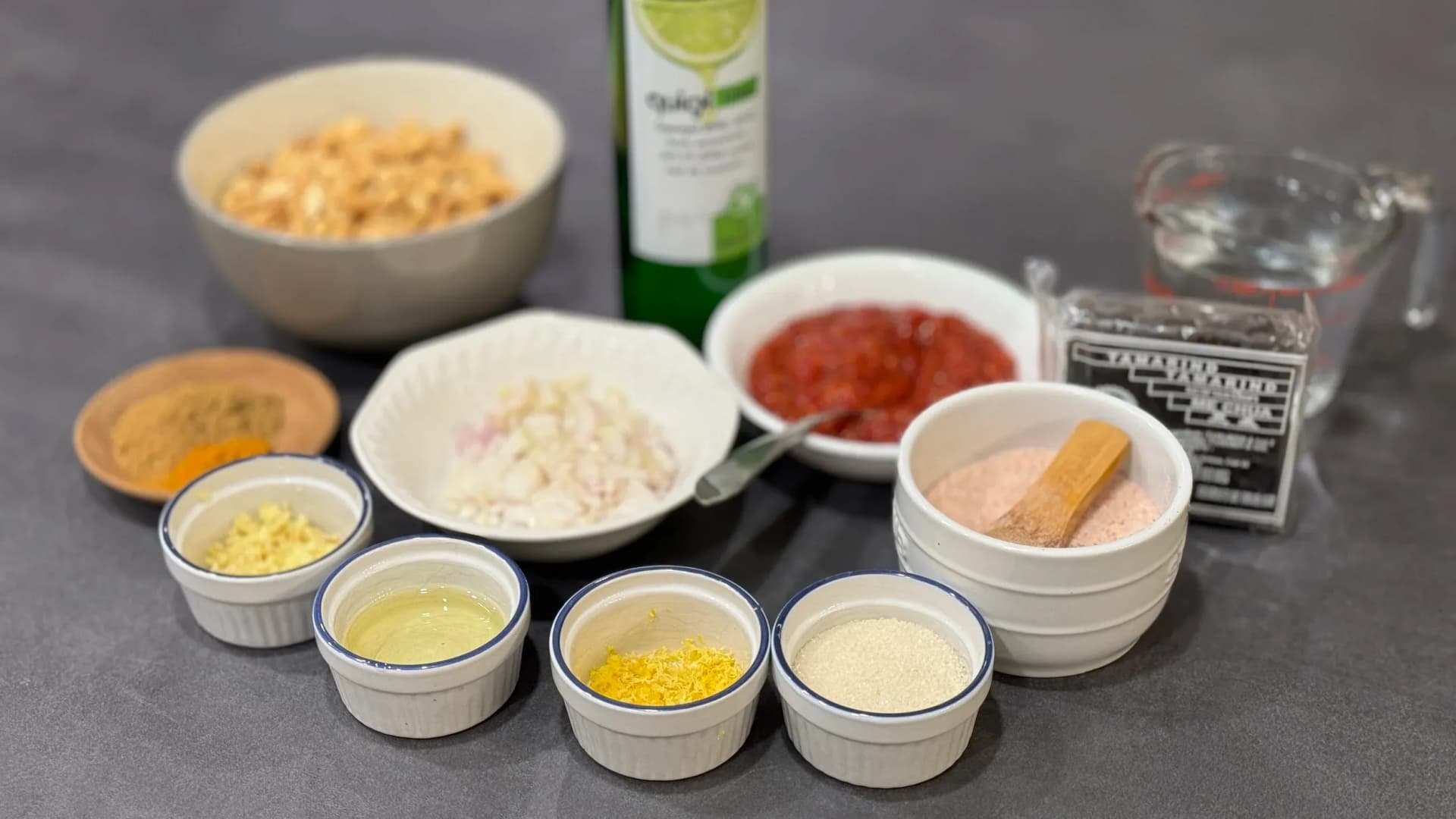 Ingredients for Singapore Chicken Satay laid out in bowls and ramekins on a gray surface, including spices, oil, and peanut sauce components.