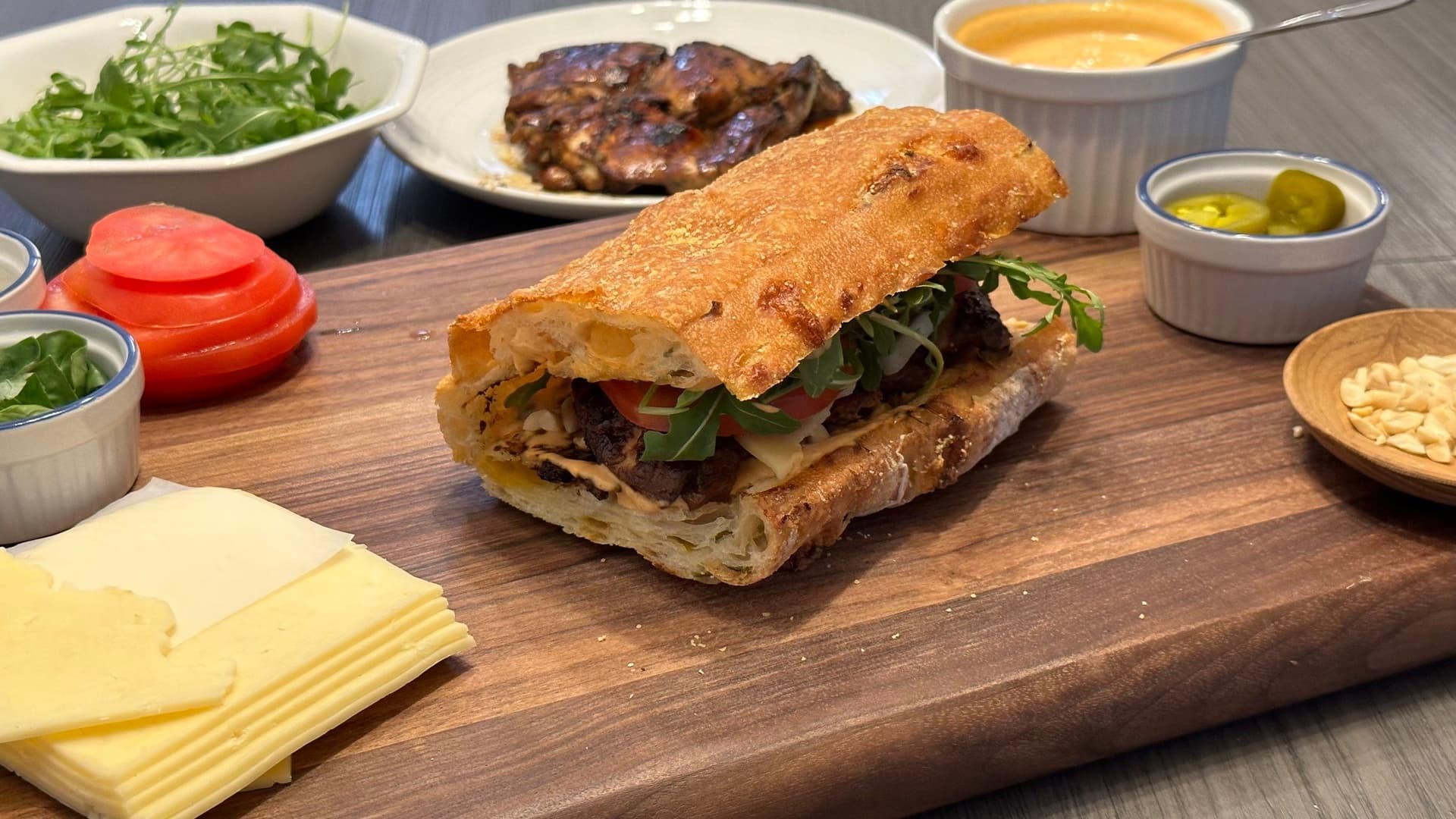 Thai chicken sandwich on wooden board with cheese, fresh greens, and condiments in bowls nearby.