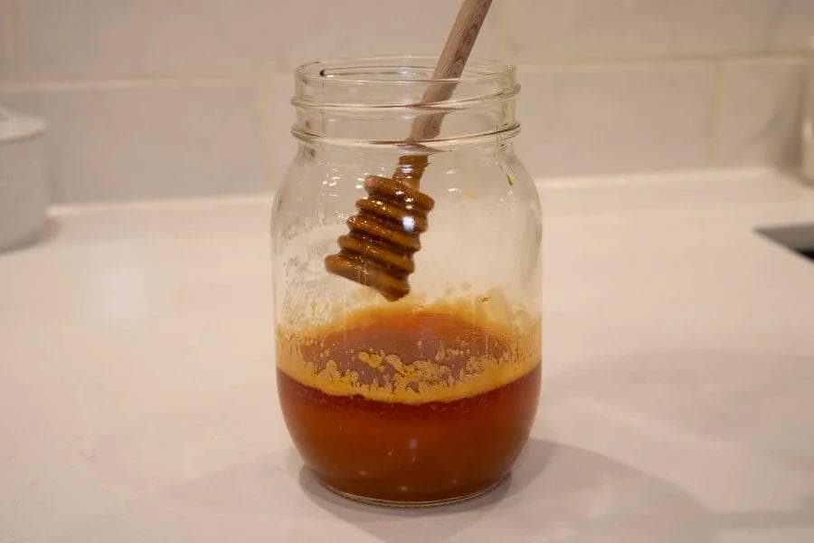 Hot honey sauce in a mason jar with a honey dipper, showing golden-brown liquid with foam on top.