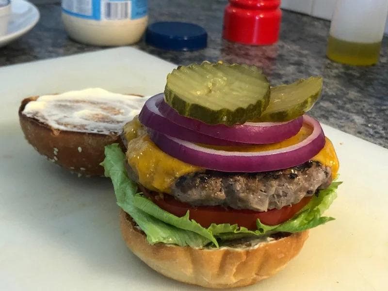 Assembled cheeseburger with lettuce, tomato, red onion, melted cheese, and pickle on a sesame seed bun with side bun.