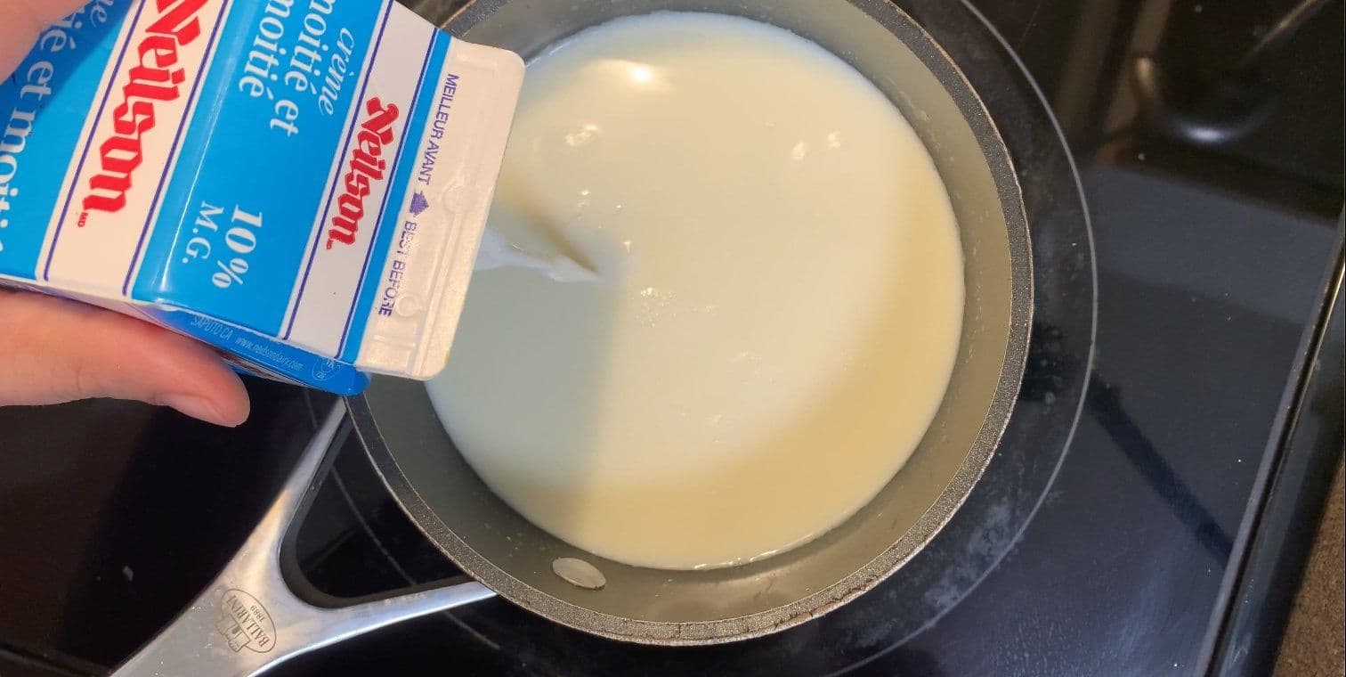Milk being poured from a carton into a saucepan containing melted white chocolate.