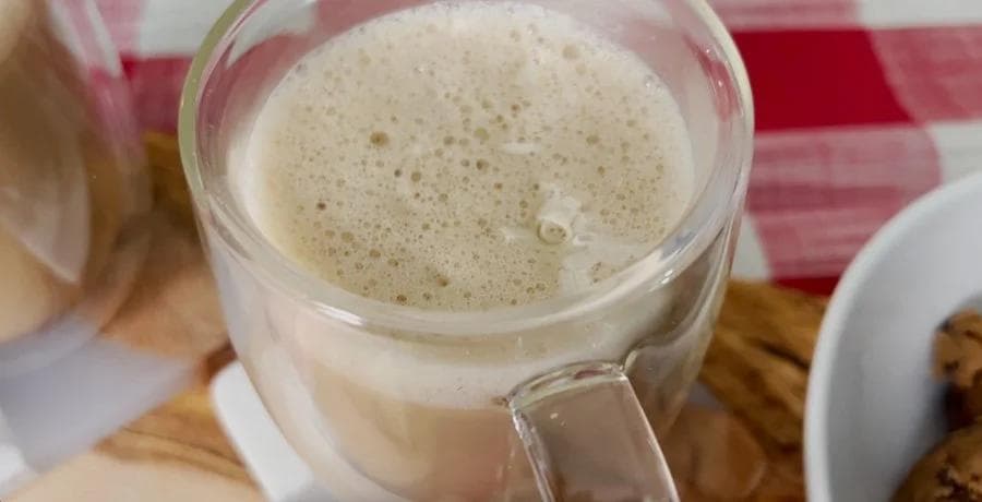 Frothy white chocolate mocha in a glass with foam top, served on a wooden surface with pink checkered cloth background.