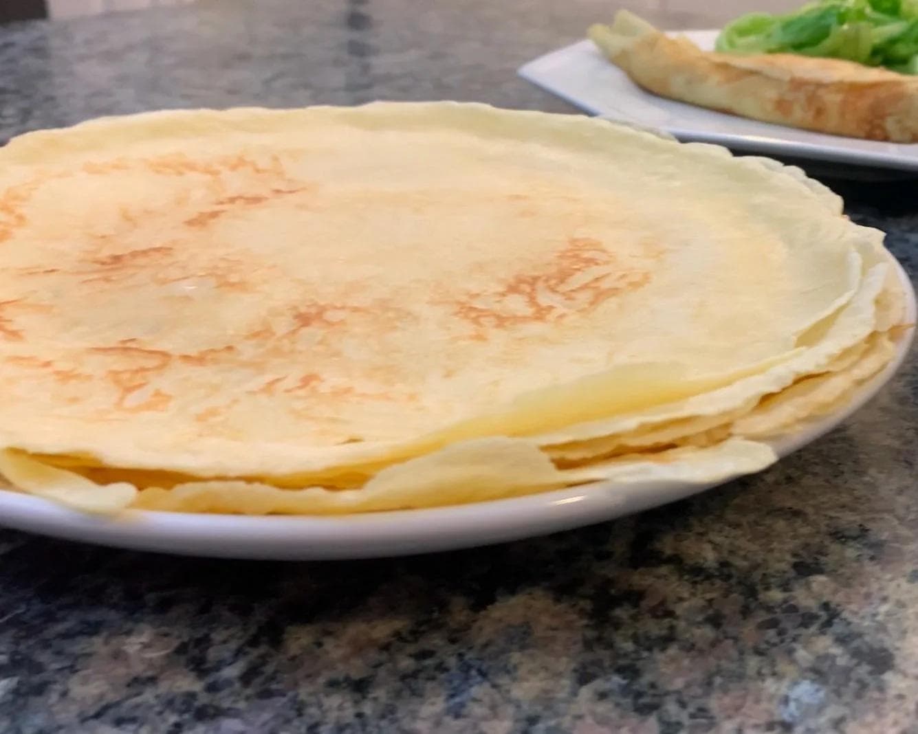 Stack of thin, golden crepes on a white plate with a gray speckled countertop background.