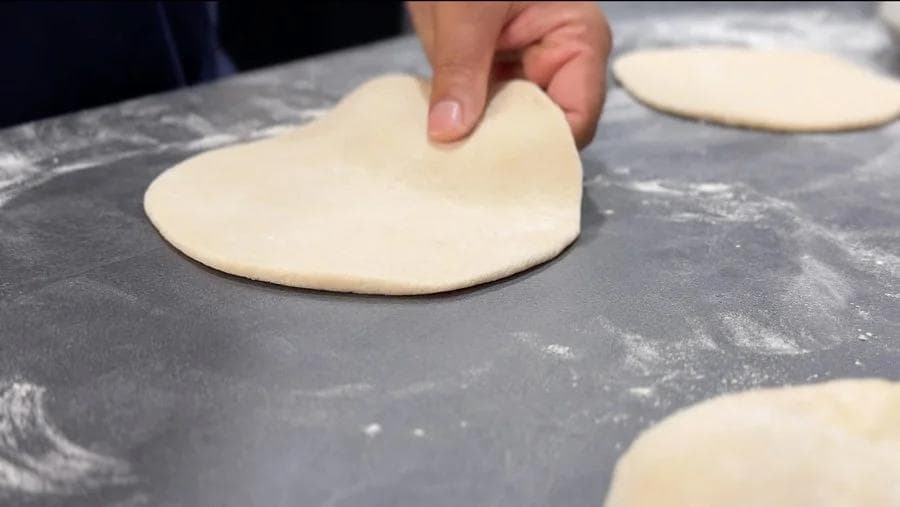 Hand flattening heart-shaped pita dough on a floured dark surface.