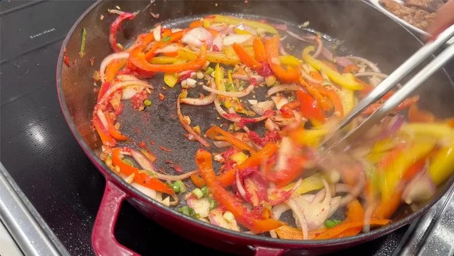 Sautéed bell peppers, onions, and aromatics sizzling in a burgundy cast iron skillet during Jamaican pepper steak preparation.