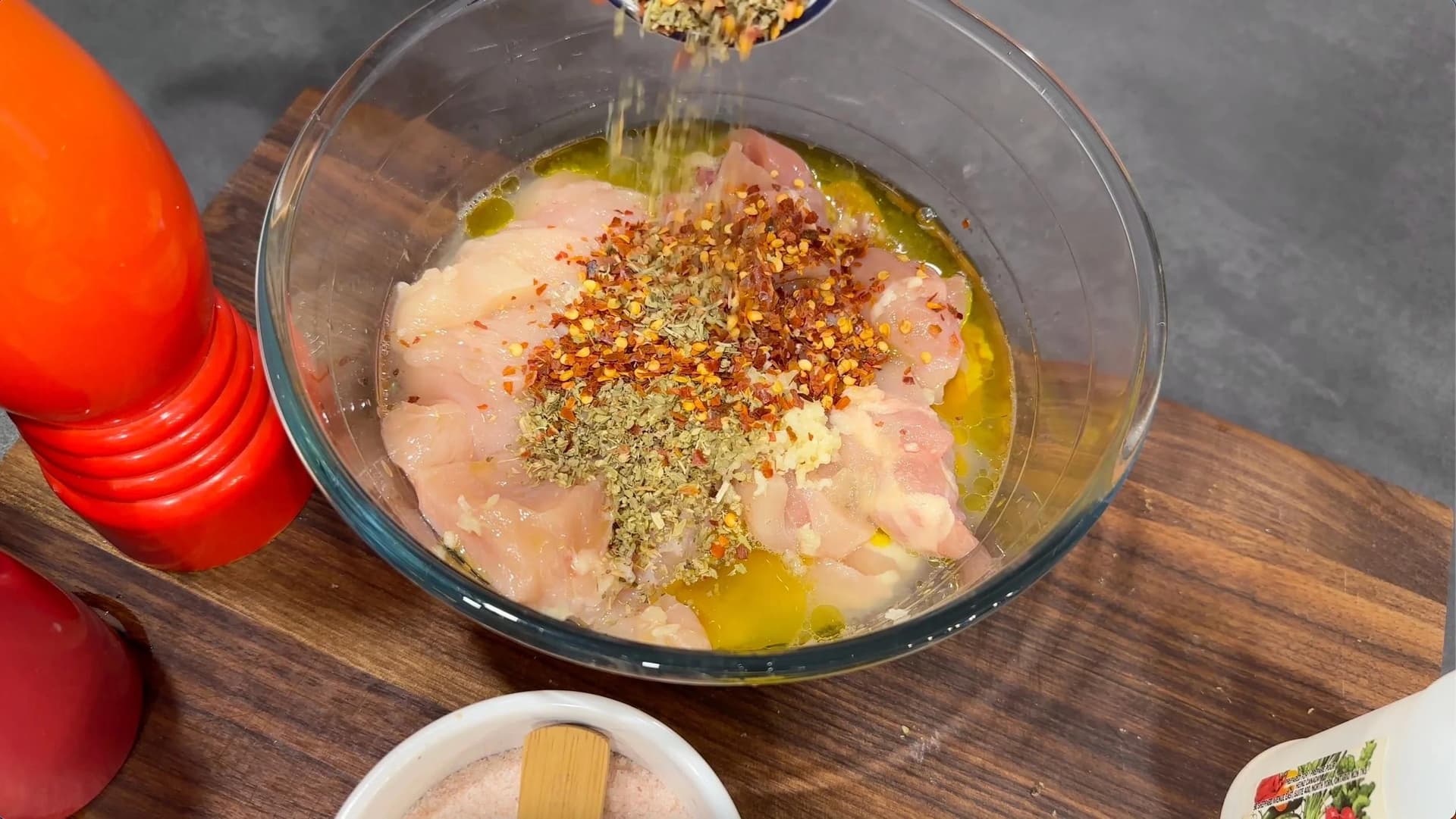 Raw chicken cubes marinating in olive oil and spices in a glass bowl with oregano and chili flakes being sprinkled on top.