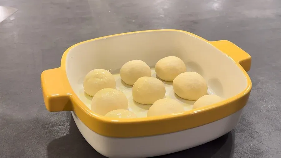 Proofed dinner roll dough balls in a yellow and white casserole dish with olive oil.
