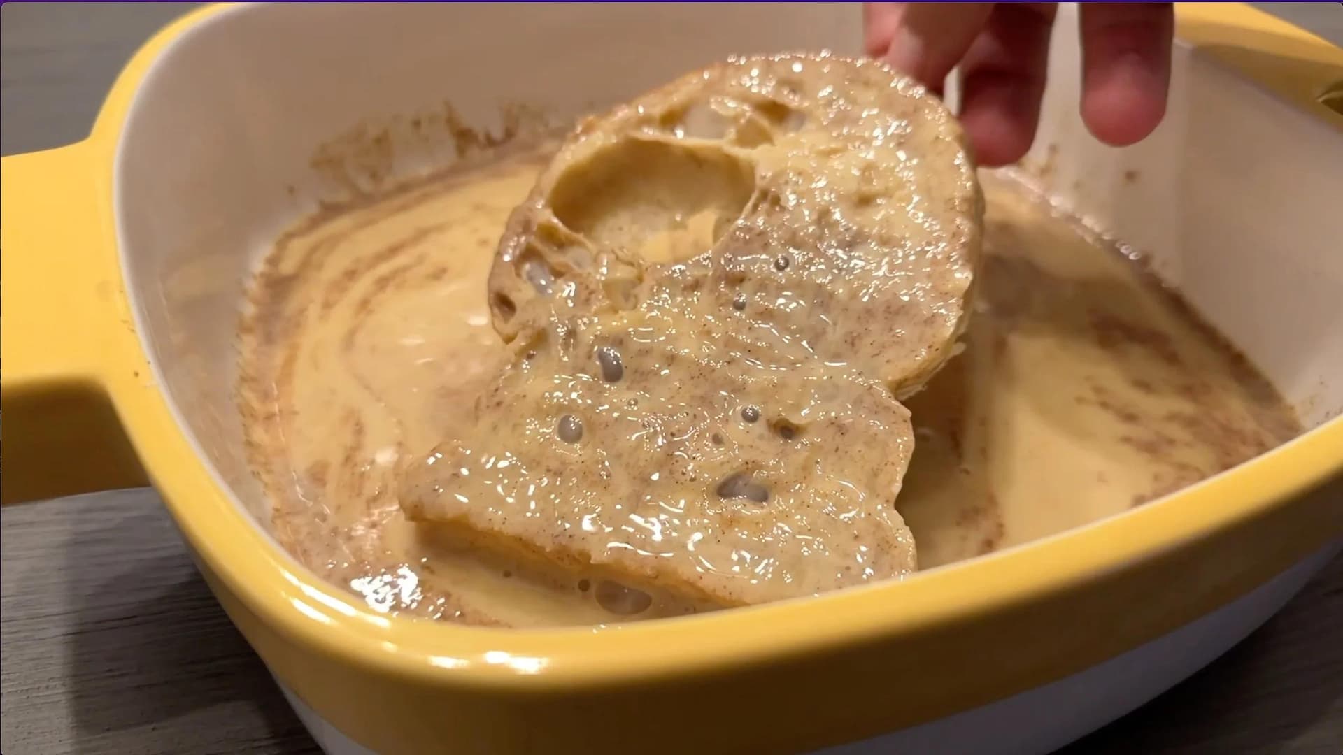 Bread slice dipped in egg mixture being lifted from yellow casserole dish for French toast preparation.