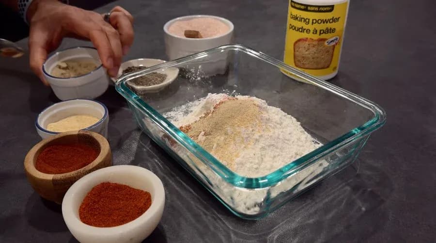 Dry ingredients being added to a glass baking dish: flour, baking powder, garlic powder, paprika, cayenne, and spices in small bowls nearby.