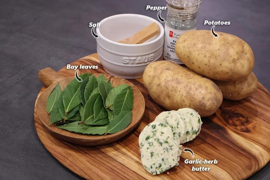 Ingredients for air fryer hasselback potatoes arranged on a wooden board: russet potatoes, garlic-herb butter, bay leaves, salt, pepper, and water.