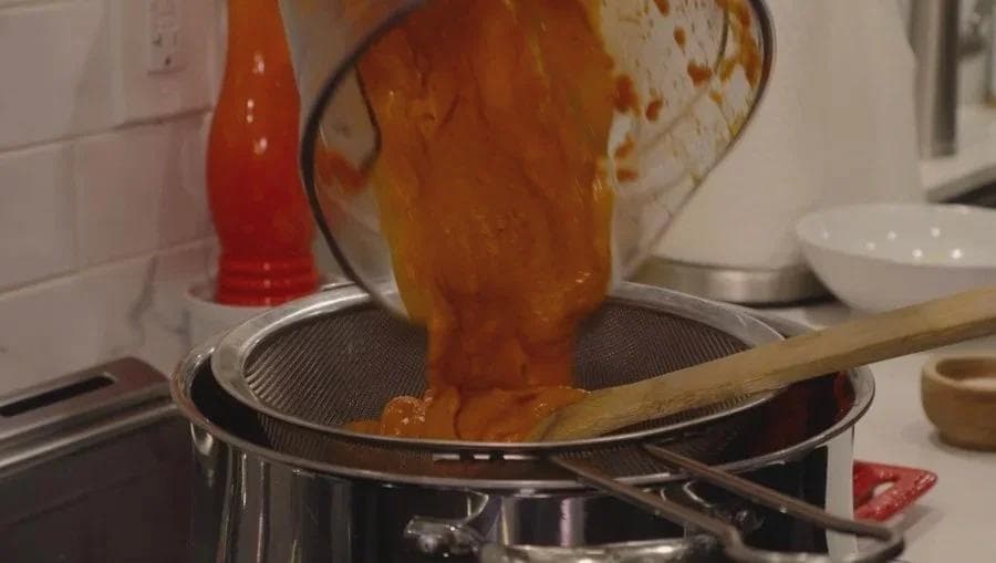 Pouring pureed butter chicken sauce through a fine mesh sieve into a pot with a wooden spoon.