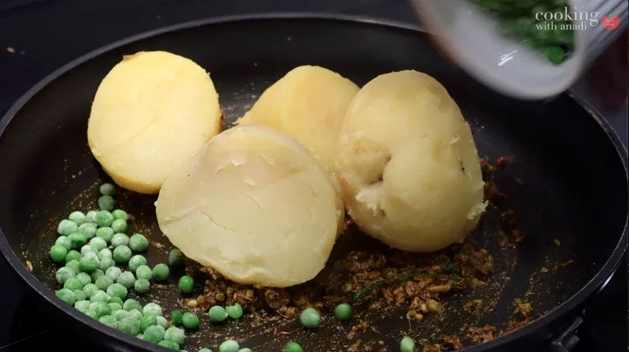 Boiled potatoes and peas being stirred into spiced curry base in a black pan.
