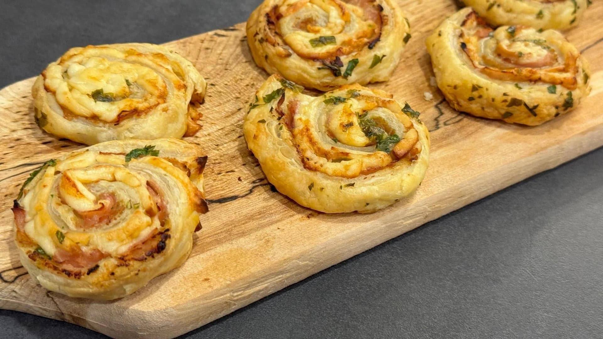 Baked ham and cheese puff pastry pinwheels garnished with fresh herbs on a wooden board.