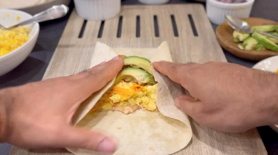 Hands holding a breakfast burrito filled with scrambled eggs, cheese, and cucumber slices wrapped in a tortilla.