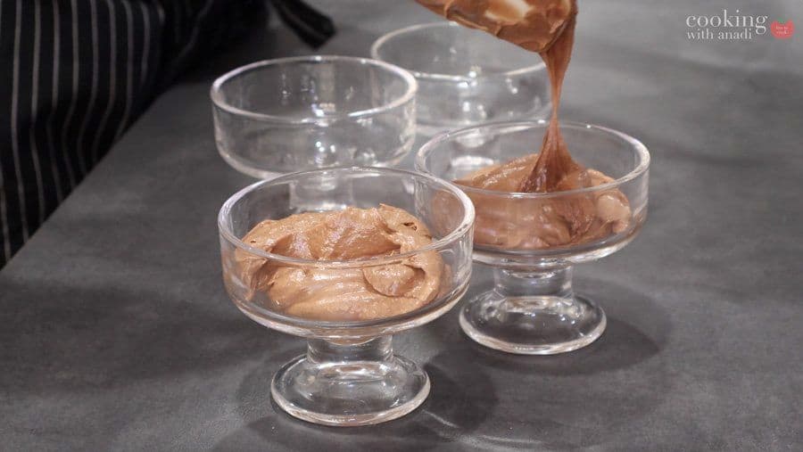 Chocolate mousse spooned into three clear glass dessert cups, with one being filled from above.