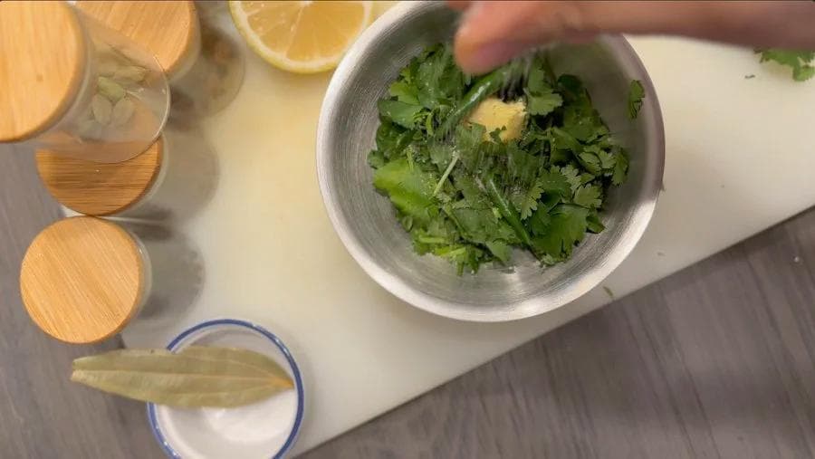 Cilantro paste being ground in a mortar with pestle, surrounded by lemon slices and other ingredients on a white countertop.