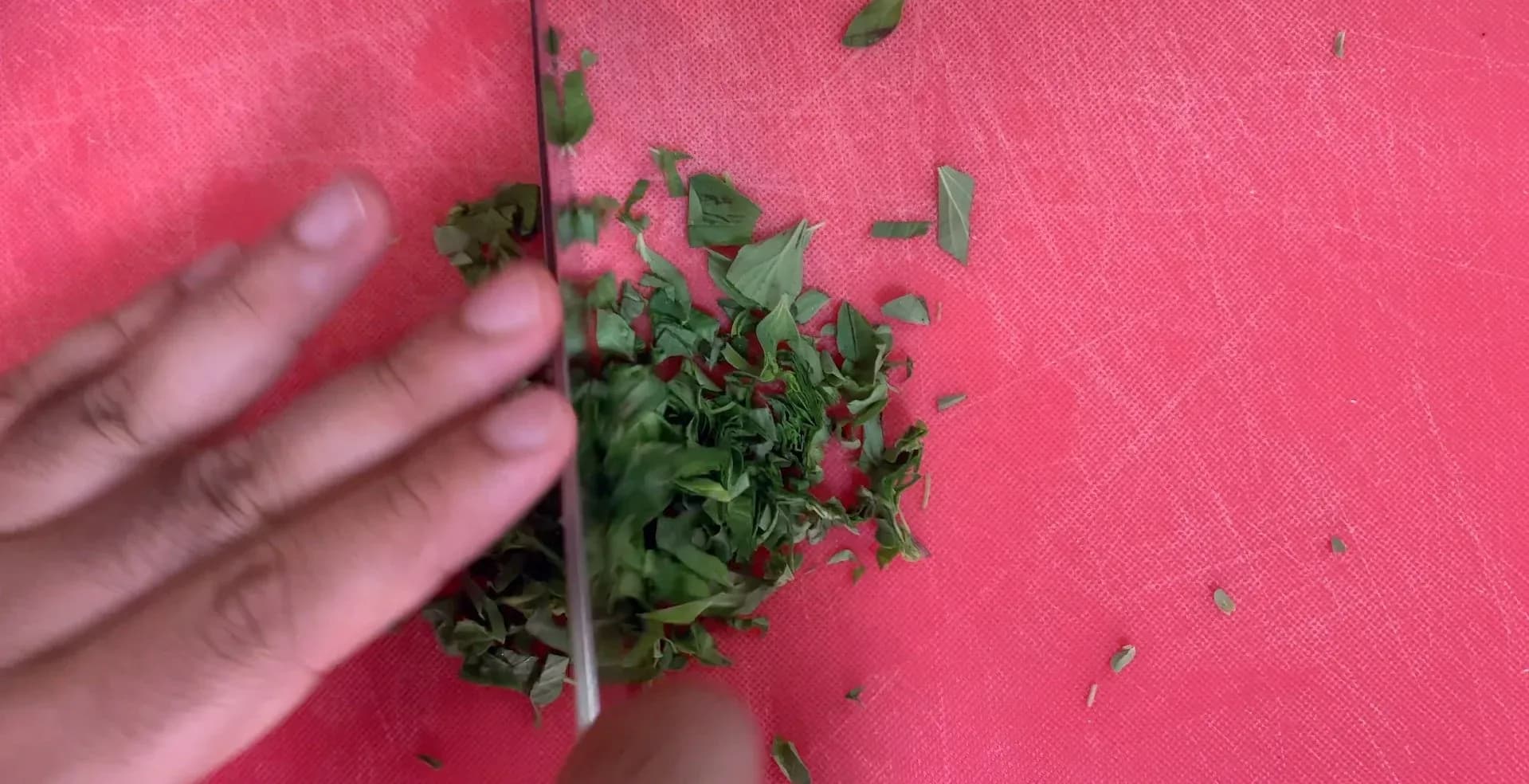 Fresh oregano leaves being chopped on a pink cutting board with a knife.