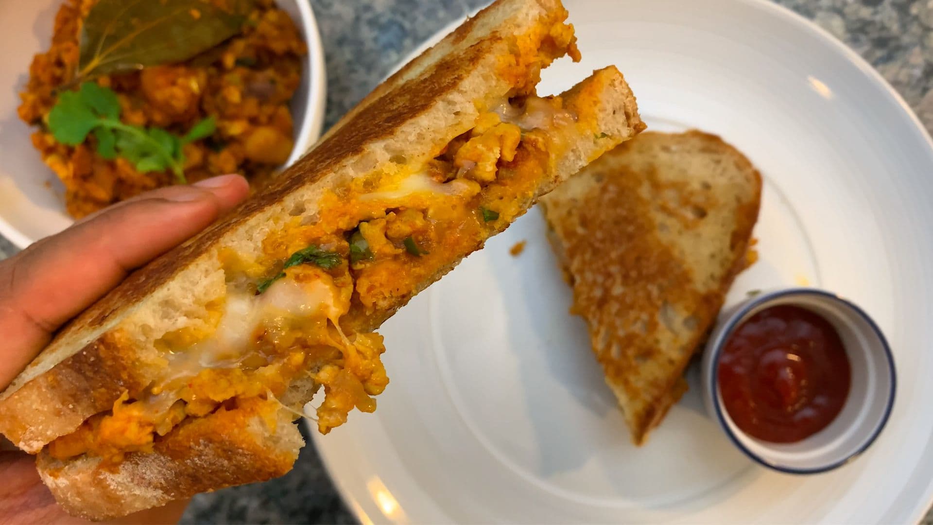 Hand holding a toasted sandwich filled with spiced chicken keema, served with ketchup and additional keema in bowls.