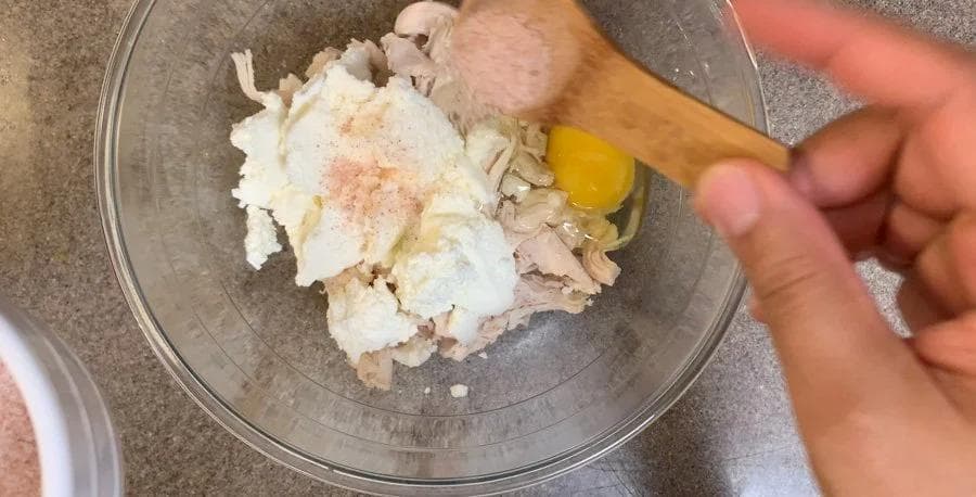 Ricotta, shredded chicken, and egg mixed in a glass bowl with a wooden spoon, seasoned with salt and pepper.