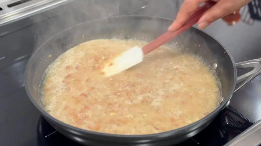 Mashing cooked beans in a skillet with broth, using a spatula to break them down into a creamy consistency.