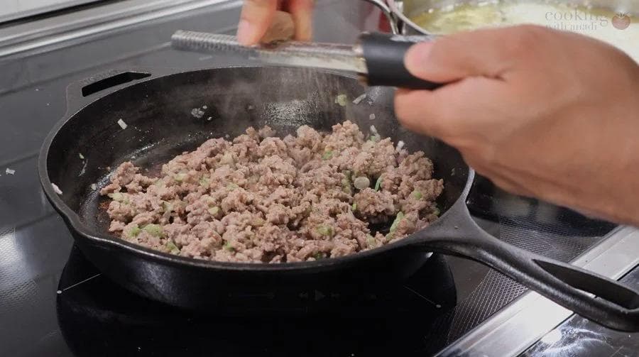 Ground meat mixture being stirred in a cast iron skillet on a stovetop during preparation of perkedel daging.