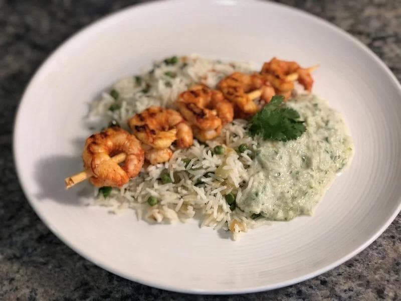 Grilled tandoori shrimp skewer served over vegetable rice pulao with green herb sauce and fresh cilantro on a white plate.