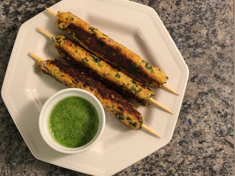 Three cooked chicken seekh kebabs on a white plate with green cilantro chutney in a small bowl