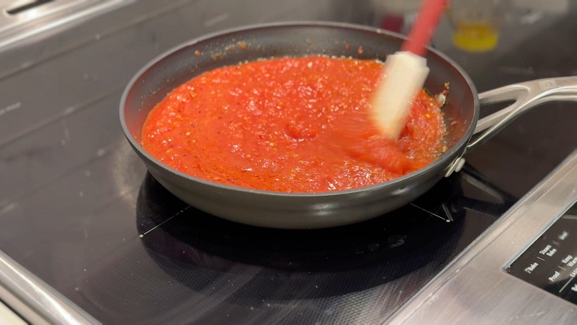 Red tomato sauce simmering in a pan on an induction cooktop with a wooden spoon stirring it.