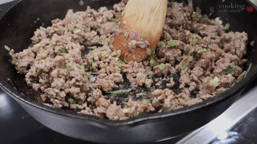 Ground beef and pork mixture being stirred in a dark skillet with a wooden spoon, mixed with minced garlic and green onions.
