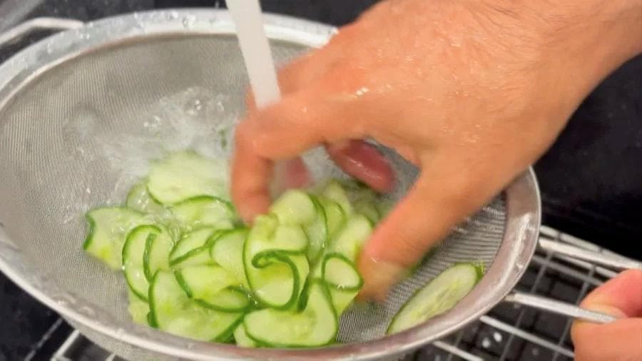 Squeezing excess water from thinly sliced cucumbers in a colander after salting.