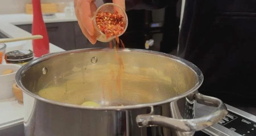 Red chilli flakes being sprinkled from a spoon into a pot of oil with onions and aromatics.