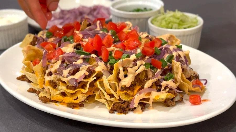 Finished cheeseburger nachos on a white plate topped with melted cheese, red tomatoes, purple onions, and sauce with condiments nearby.