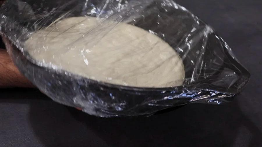 Pizza dough covered with plastic wrap proofing in an oiled cast iron pan.