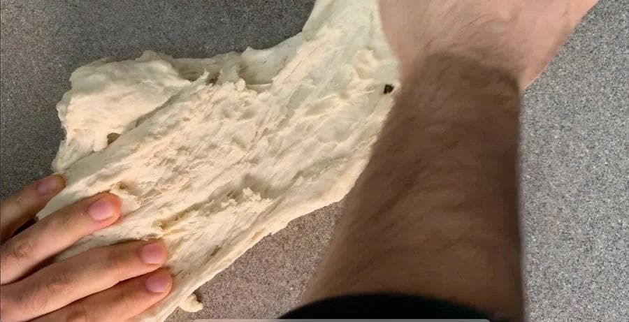 Hands stretching and kneading pizza dough on a gray countertop during the folding and stretching step.