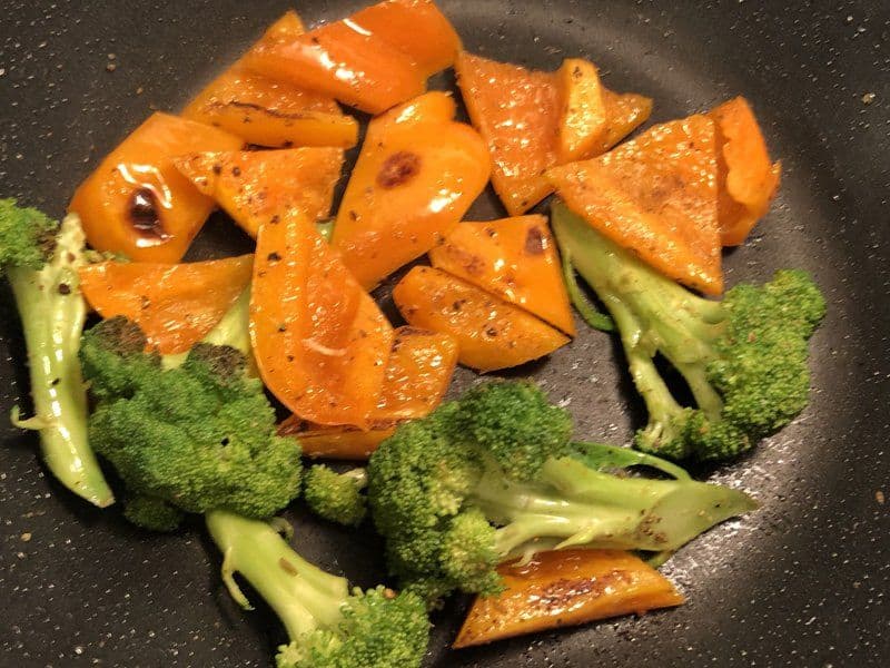 Teriyaki shrimp stir fry with broccoli florets and orange bell pepper slices cooking in a dark pan.