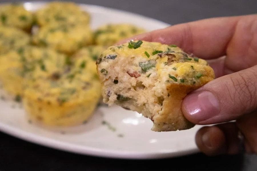 Hand holding a bite-sized egg muffin showing fluffy interior with herbs and meat pieces visible.