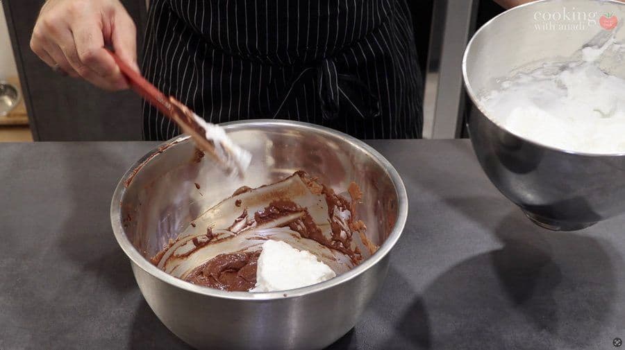 Folding cooled chocolate into egg yolk mixture with a spatula in a metal bowl during mousse preparation.