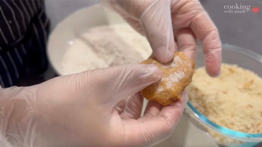 Hand pressing breaded chicken nugget into breadcrumb coating in glass bowl.