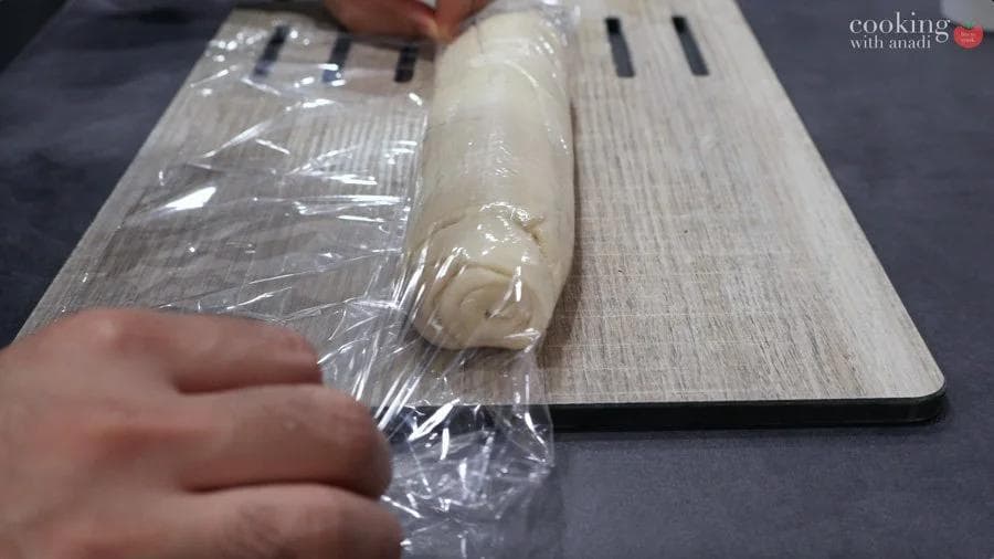 Crescent dough rolled with mashed potatoes wrapped in plastic wrap on a wooden cutting board before chilling.